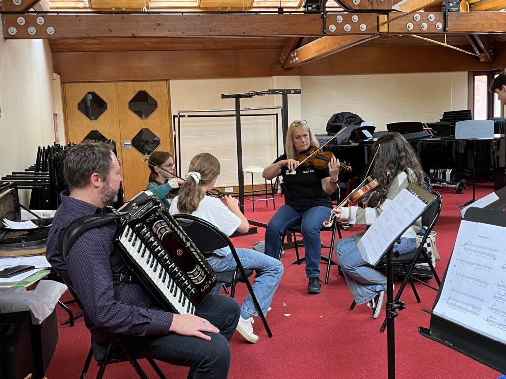 A group of students playing violins sat working with two tutors, one playing an accordian and another playing a violin.